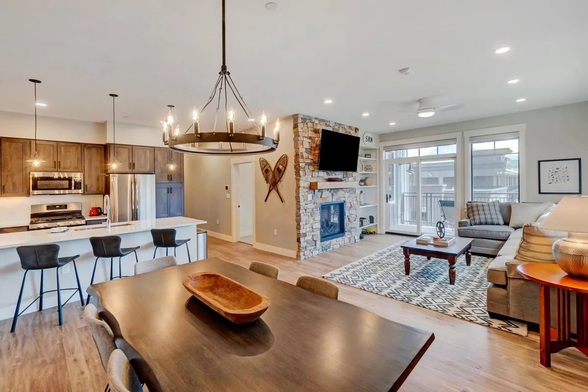 The image showcases a spacious, open-concept living and dining area with a cozy fireplace, a modern kitchen island, a stylish dining table set for a meal, and a comfortable seating area perfect for gathering. The ambiance is warm and inviting, ideal for relaxing after a day of mountain adventures.