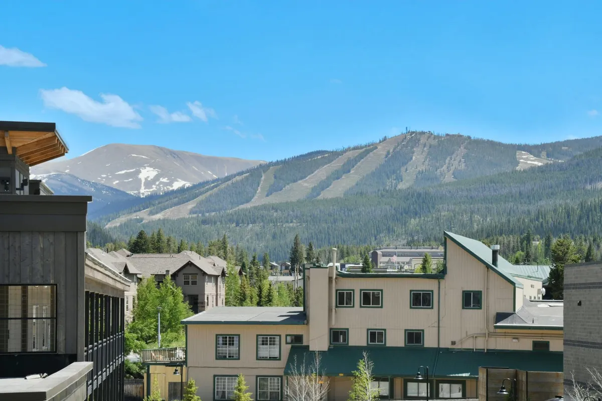A panoramic view blending modern mountain architecture with an expansive mountain landscape under a clear blue sky. The scene suggests proximity to hiking and skiing trails and gives a sense of the majestic outdoor environment surrounding the property.