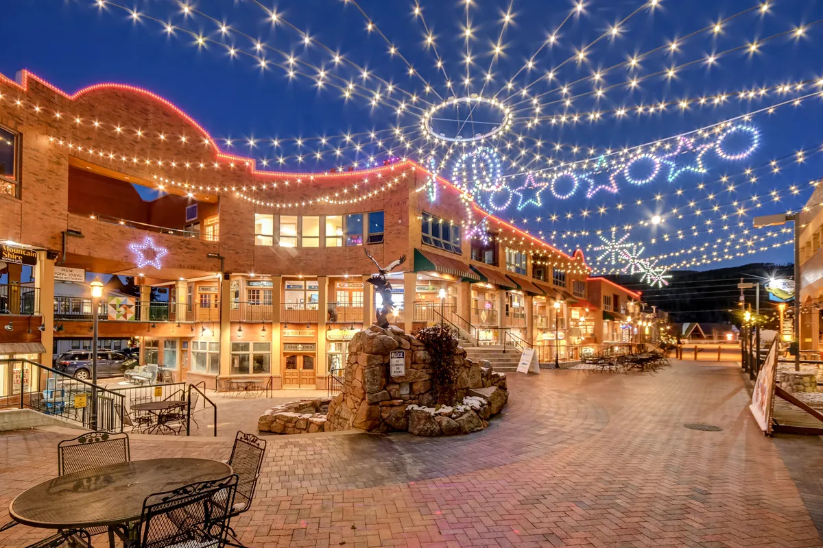 Twinkling lights hang overhead in an outdoor plaza adorned with decorative stars and a stone fountain at its center. The setting is festive, with people engaging in evening strolls, outdoor dining, and perhaps attending holiday events or community gatherings. The atmosphere is joyful, warm, and inviting, capturing the vibrancy of local life.