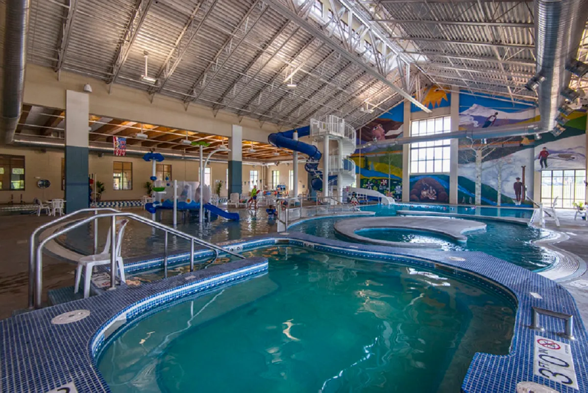 A vibrant indoor pool area featuring thrilling water slides, sparkling swimming pools, colorful murals, and plenty of lounge chairs for relaxing. Families and children are seen enjoying water games and relaxation by the poolside, creating an energetic and playful atmosphere.