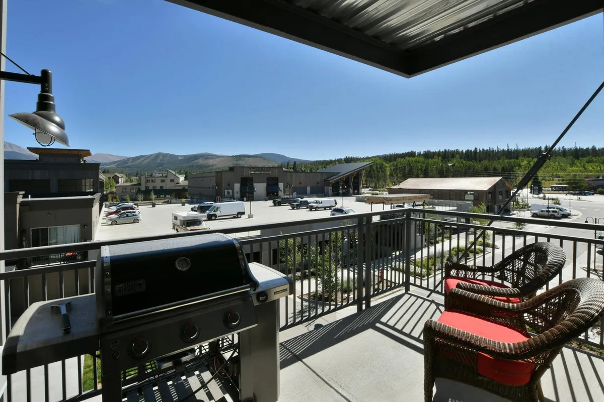 A cozy outdoor balcony featuring a gas grill and comfortable seating, set against a stunning backdrop of mountains. The scene is perfect for outdoor dining, relaxing with friends, or simply soaking in the view. Warm, inviting lighting hints at the possibility of both sunrise coffee moments and evening gatherings.