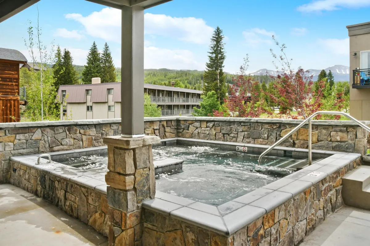 An inviting exterior shot featuring a large, bubbling hot tub set against a stunning mountain landscape. The hot tub is surrounded by a spacious deck that offers scenic views, perfect for relaxing and socializing after a day of outdoor adventure.